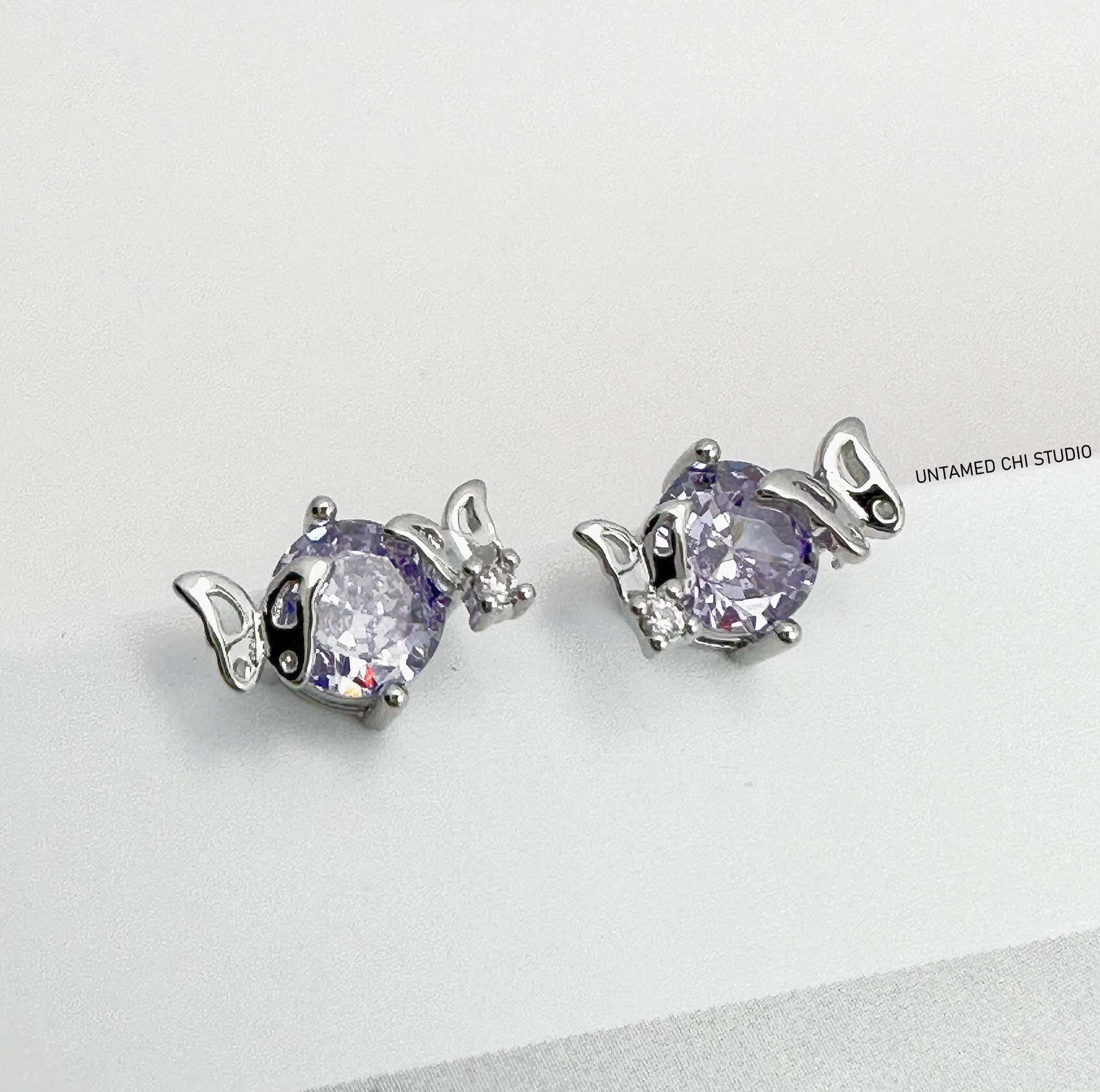 Elegant oval-cut lavender stud earrings encased in silver settings with petite wing-like designs on the sides, displayed against a white backdrop with 'UNTAMED CHI STUDIO' text