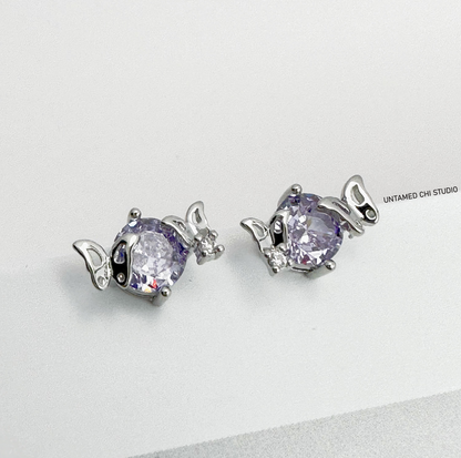 Elegant oval-cut lavender stud earrings encased in silver settings with petite wing-like designs on the sides, displayed against a white backdrop with 'UNTAMED CHI STUDIO' text
