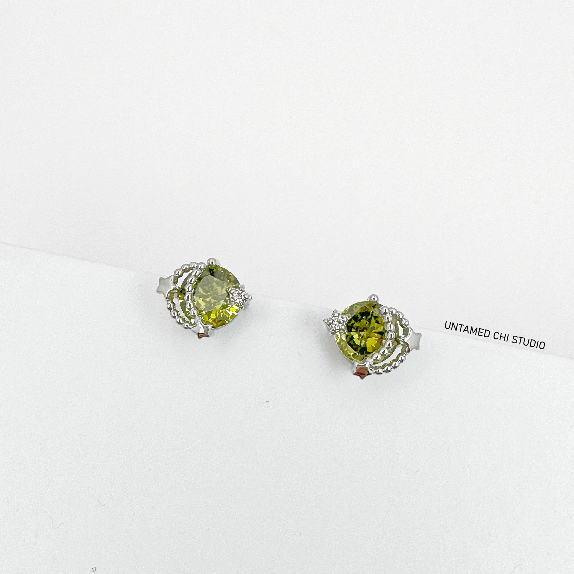 Green gemstone stud earrings with a faceted cut, surrounded by a halo of tiny white gems set in a silver finish, on a white display stand with 'UNTAMED CHI STUDIO' visible.